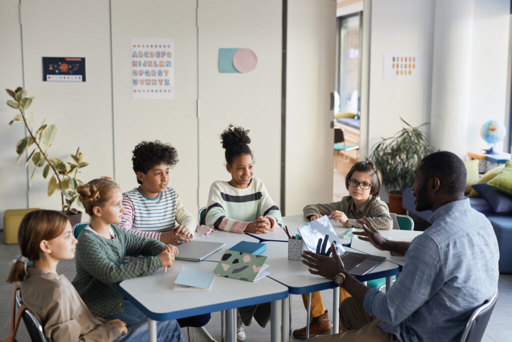 Diverse group of children sitting at table with male teacher in modern school classroom